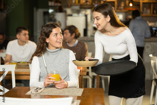Female waitress brings freshly cooked food to a client against the background of other visitors to the restaurant. European woman gets quality food and service in a restaurant