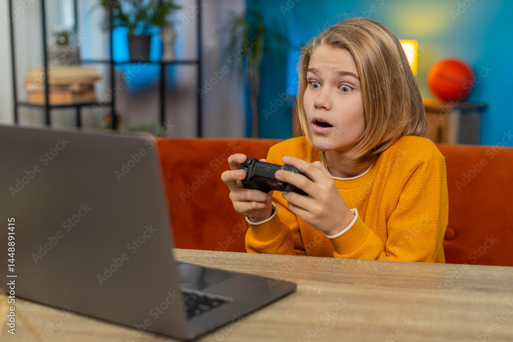 Caucasian teenager boy playing a football sports simulator on laptop at home. Child winner using a joystick to compete, joyfully celebrating a scored goal and victory while sitting on couch by table Caucasian teenager boy playing a football sports simulator on laptop at home. Child winner using a joystick to compete, joyfully celebrating a scored goal and victory while sitting on couch by table
