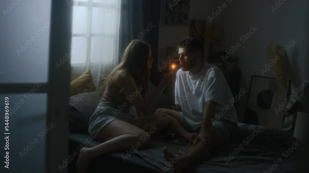 Through-doorway full shot of young couple resting in bed in homewear, lady lighting cigarette for beloved boyfriend who smoking indoors, affectionate love and peacefullness Through-doorway full shot of young couple resting in bed in homewear, lady lighting cigarette for beloved boyfriend who smoking indoors, affectionate love and peacefullness