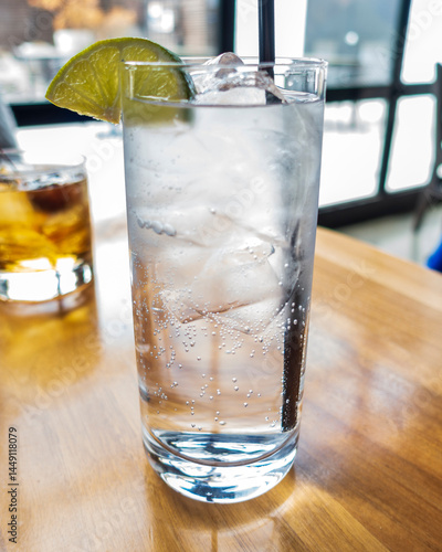 Obraz na płótnie A crisp gin and tonic served in a tall glass packed with ice, topped with a lime wedge. Tiny bubbles rise through the clear drink, catching the sunlight on a wooden table by a bright window. w salonie