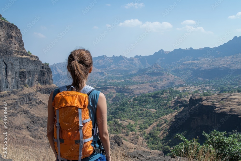 Naklejka premium Woman with orange backpack looks out over a vast, arid mountain range. Use this photo to illustrate travel, adventure, and seeing the world.
