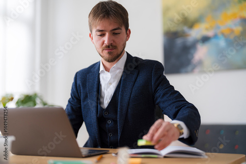 A businessman in a suit works at his desk. He is using a highlighter on a notebook while his laptop is open.