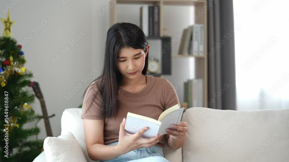 Young woman sitting on sofa, reading book in cozy living room with Christmas tree, modern decor, warm light, relaxation and peaceful atmosphere Young woman sitting on sofa, reading book in cozy living room with Christmas tree, modern decor, warm light, relaxation and peaceful atmosphere