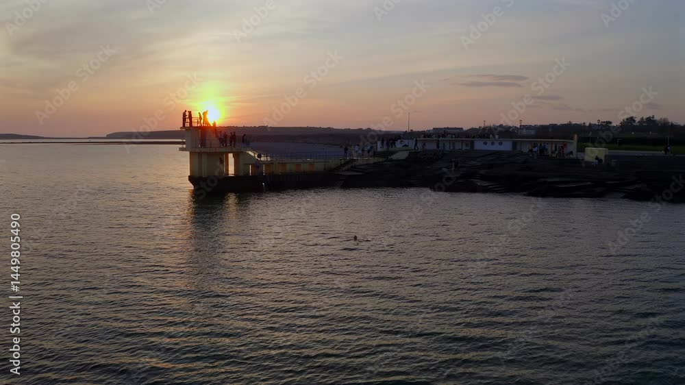 Aerial establishing of pier and tower as light fades and golden hues wash across calm sea as people swim and stand along railing Aerial establishing of pier and tower as light fades and golden hues wash across calm sea as people swim and stand along railing