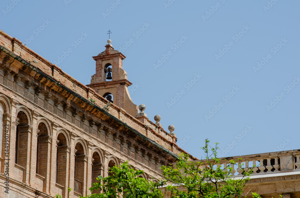 Obraz premium Elegant architectural detail of an old European church with a classic bell tower, surrounded by clear blue sky and green tree branches, perfect for travel and history themes.