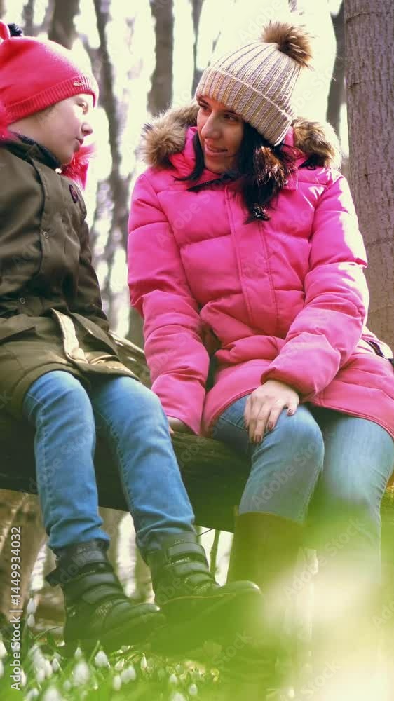In the spring forest, among the snowdrops, a young woman and a teenager girl are sitting on a log, a large branch of a fallen tree, talking. in the afternoon. Happy family in the spring forest In the spring forest, among the snowdrops, a young woman and a teenager girl are sitting on a log, a large branch of a fallen tree, talking. in the afternoon. Happy family in the spring forest
