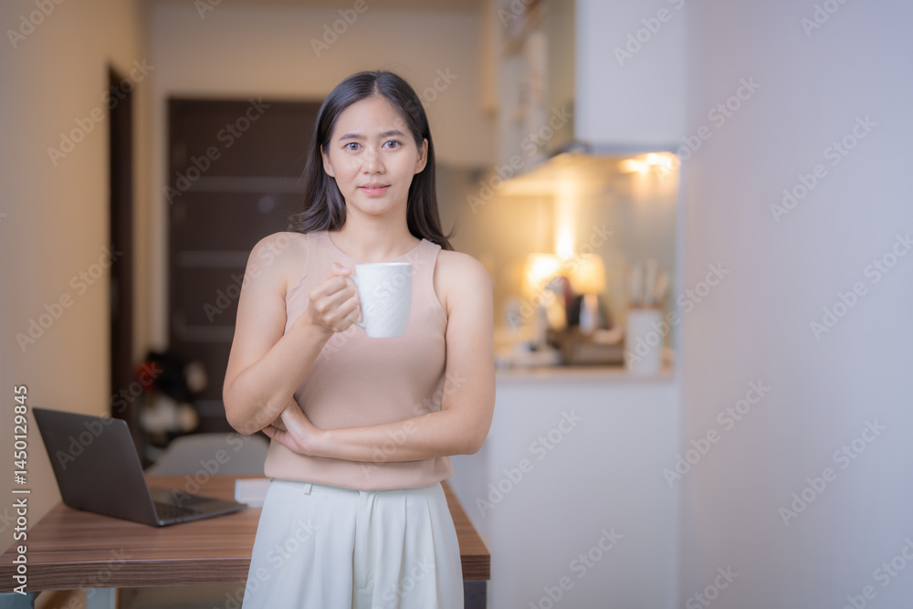 Young Asian woman standing in a modern kitchen, holding a white mug while working from home. Laptop resting on the table, creating a comfortable and relaxed atmosphere Young Asian woman standing in a modern kitchen, holding a white mug while working from home. Laptop resting on the table, creating a comfortable and relaxed atmosphere