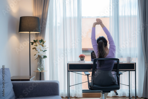 Young Asian businesswoman sitting on an ergonomic chair is stretching her arms after using a laptop computer for a long time in her modern home office
