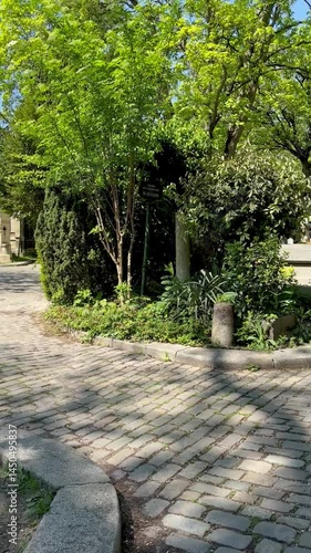 Tranquil cobblestone path winding through historic mausoleums at Père Lachaise Cemetery in Paris, France, surrounded by lush spring greenery under clear blue skies.