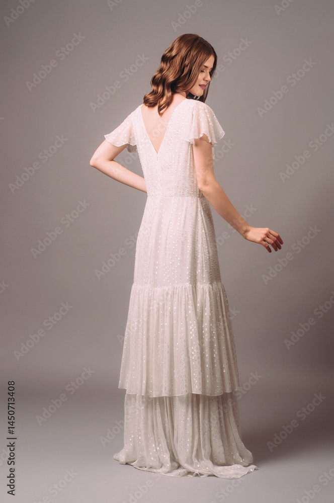 Fototapeta premium White shiny maxi gown with tiered skirt. Romantic portrait of smiling brunette woman in bohemian evening dress with short sleeves. Bridesmaid's fashion. Summer feminine look for hen party, rear view