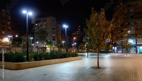 Nighttime urban plaza with illuminated buildings and trees.  Well-lit paved area, low-level landscaping, and modern streetlights