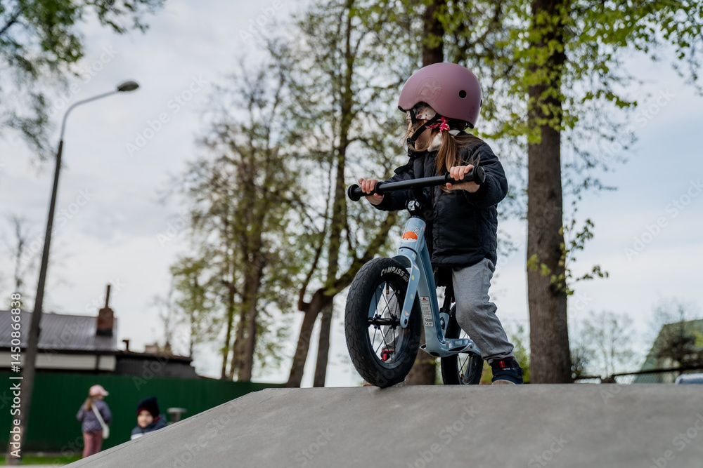 Obraz premium Small little girl with pink protective head gear helmet on small blue pedal-less balance bike riding on a track for learning new riding skills