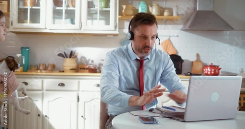 Cheerful little girl plays with her dog and distracts her business father, who working remotely from home, having an important business video conference call on laptop in the kitchen.
