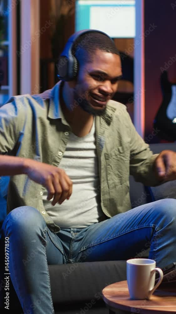 Vertical Video African American guy working on laptop celebrating achievement under neon lights, sitting on a couch in cozy apartment. Relaxed person listens to music, pleased with tasks success Vertical Video African American guy working on laptop celebrating achievement under neon lights, sitting on a couch in cozy apartment. Relaxed person listens to music, pleased with tasks success
