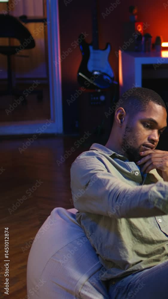 Vertical Video Black man taking a break from work by scrolling his smartphone at home, messaging friends on social media and having fun. Guy relaxing in stylish living room with neon lights. Camera A. Vertical Video Black man taking a break from work by scrolling his smartphone at home, messaging friends on social media and having fun. Guy relaxing in stylish living room with neon lights. Camera A.
