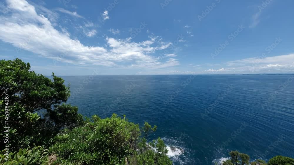 Panoramic view of tranquil blue ocean waters from lush coastal cliffs with gentle waves breaking under a clear sky filled with soft clouds Panoramic view of tranquil blue ocean waters from lush coastal cliffs with gentle waves breaking under a clear sky filled with soft clouds