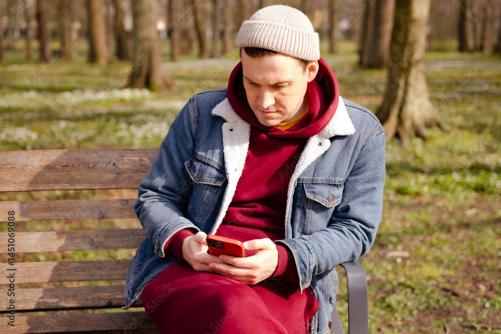 A young man dressed in a cozy red hoodie and denim jacket sits on a wooden bench in a park absorbed in his smartphone surrounded by trees on a bright day A young man dressed in a cozy red hoodie and denim jacket sits on a wooden bench in a park absorbed in his smartphone surrounded by trees on a bright day