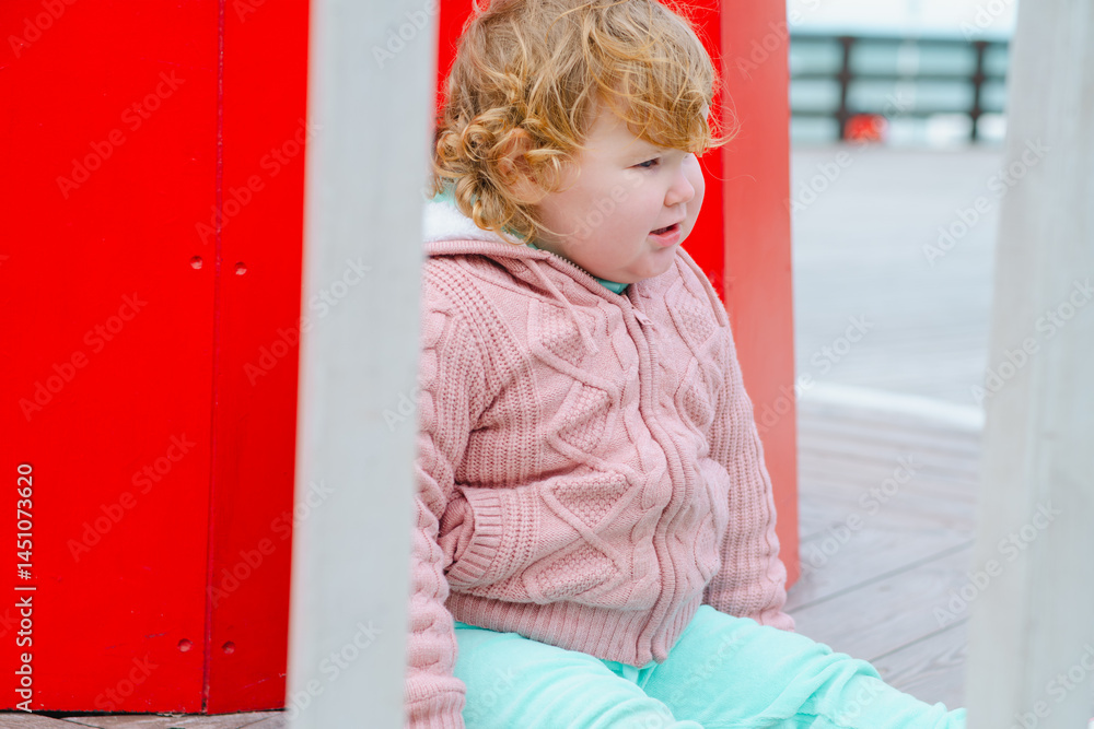 Fototapeta premium A young child dressed in a cozy sweater and light pants sits on a wooden structure at an outdoor playground The wind gently tousles her hair as she explores her surroundings