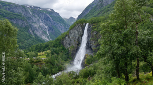 A beautiful waterfall cascades down a rocky cliff face surrounded by lush green trees and towe mountains under an overcast sky in the Norwegian wilderness.