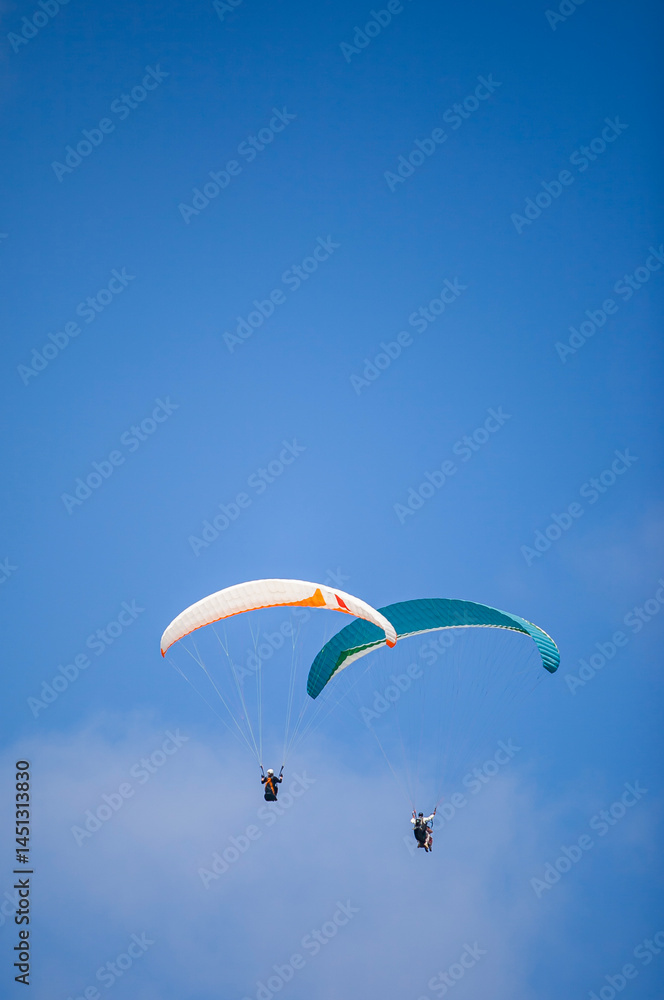 A pair of paragliding athletes are floating in the air showing their skills in controlling their paraglider against a backdrop of bright blue sky.