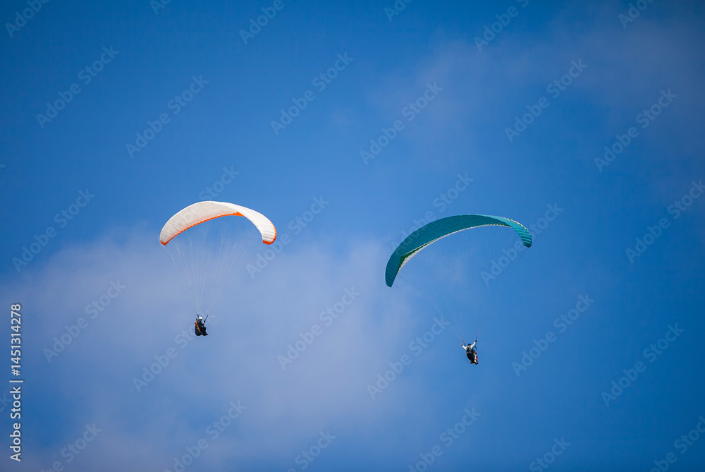 A pair of paragliding athletes are floating in the air showing their skills in controlling their paraglider against a backdrop of bright blue sky.