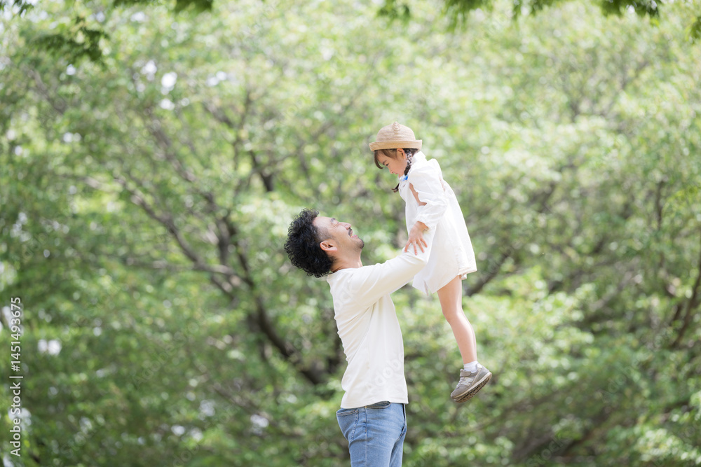 Fototapeta premium 緑の背景で公園で娘を抱っこして高い高いする父親 と娘の親子の幸せや未来、幸福のイメージ
