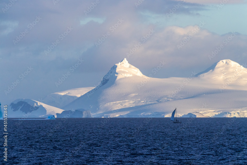 An Antarctic landscape, taken during the golden hour, just after sunrise, in the Gerlache Strait. A little sailboat in the foreground explores the Antarctic waters.