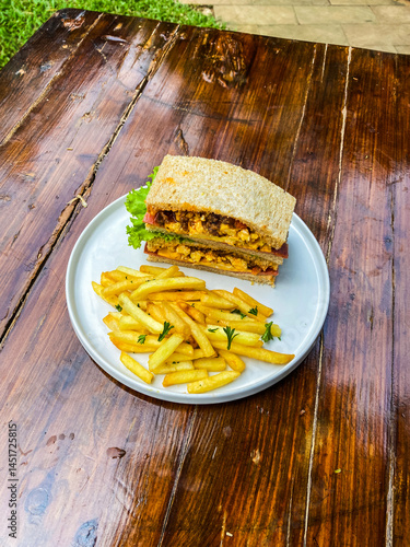 Vertical shot of a sandwich and fries on a blue rimmed plate, displayed on a wooden table in a natural outdoor setting.