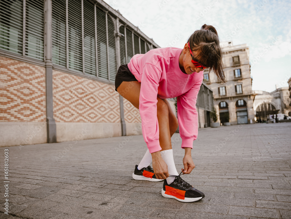 In cloudy weather in daylight, a happy dark-haired European adult woman in sunglasses in a pink raglan is tying shoelaces on a sneaker while bending down on the street. In cloudy weather in daylight, a happy dark-haired European adult woman in sunglasses in a pink raglan is tying shoelaces on a sneaker while bending down on the street.