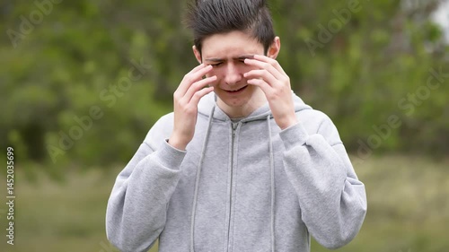 Sad depressed teenage boy crying and rubbing eyes with hands outdoors. Downcast young man wiping tears from eyes feeling overwhelmed. Sorrowful adolescent boy shedding tears and covering face with