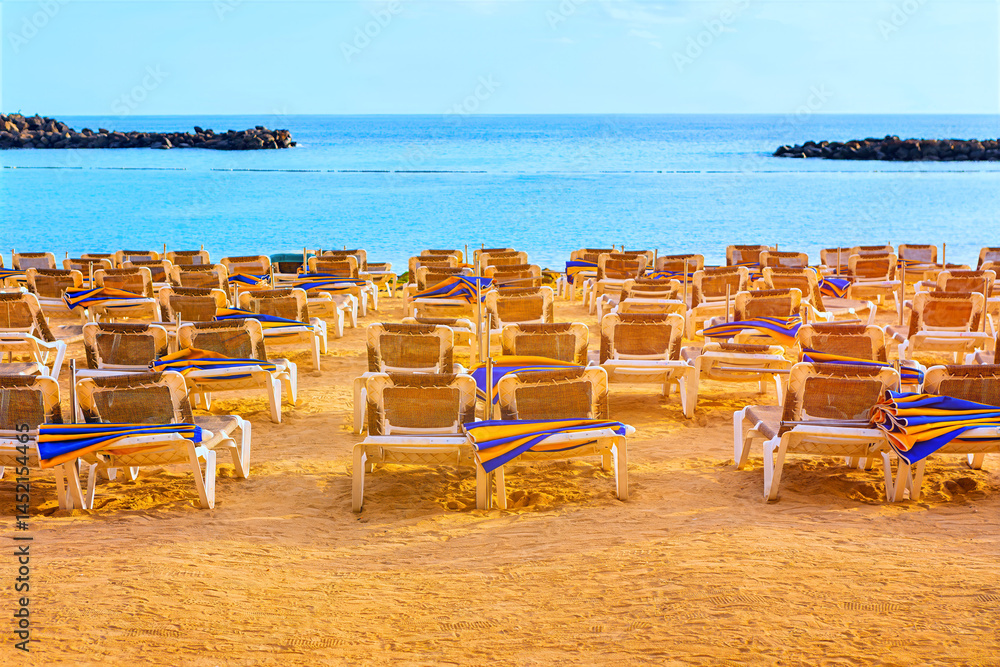 Empty beach chairs line the shore. Calm ocean breeze rustles beach umbrellas, draped over empty chairs. Rows of lounge chairs with colorful towels sit on sandy beach, facing a calm turquoise sea