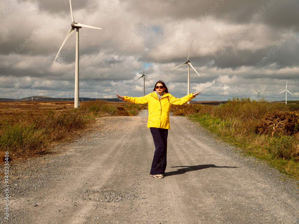 Young teenager girl in yellow jacket standing on a road with hands up, Wind turbines and vast wild fields in the background. Learning clean and green energy production. Warm sunny day.