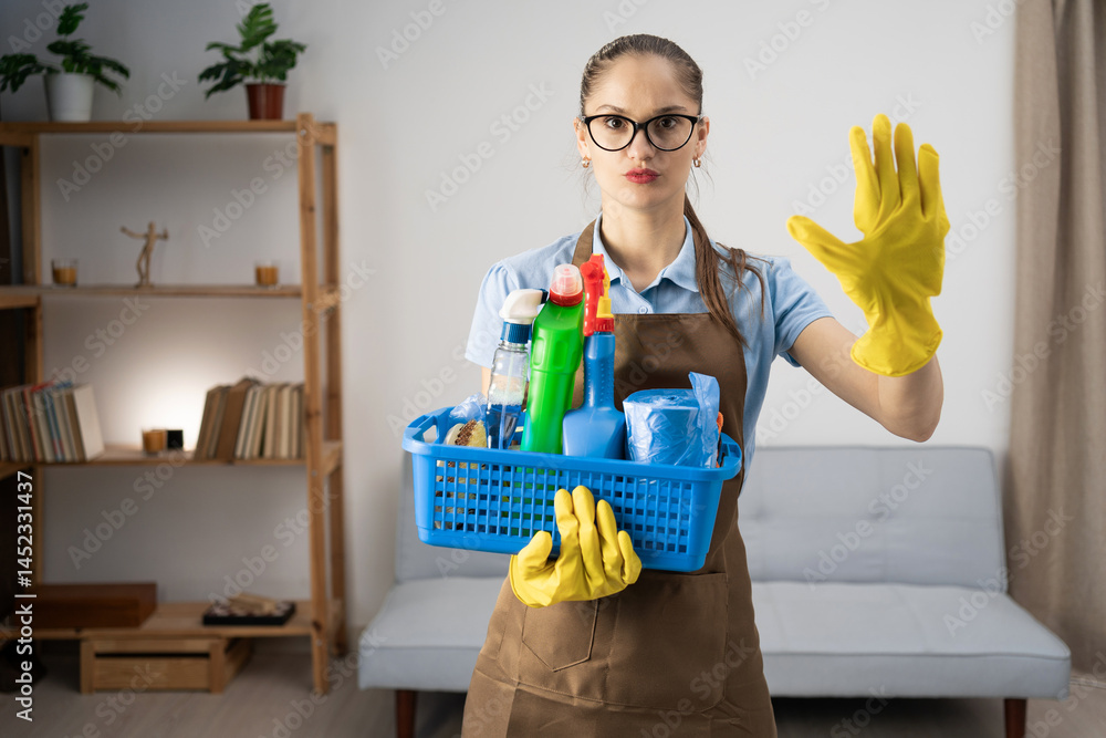 Cleaner woman wearing apron holding cleaning products in basket standing in the living room doing stop gesture with hand palm. Cleaning Cleaner woman wearing apron holding cleaning products in basket standing in the living room doing stop gesture with hand palm. Cleaning