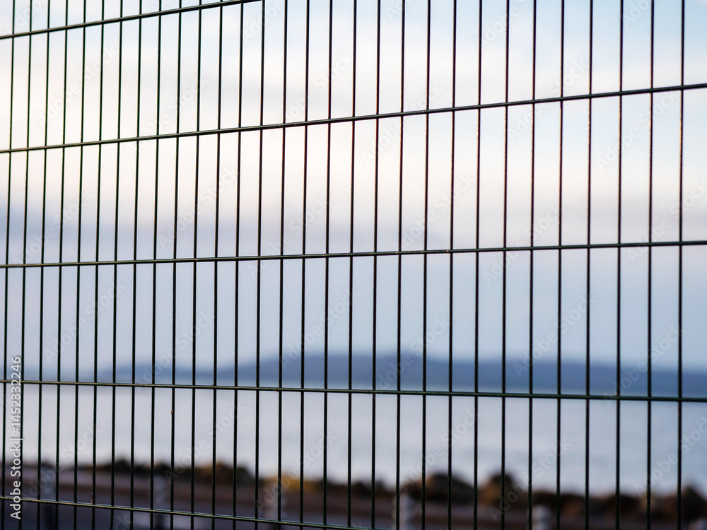 Metal wired fence in focus, ocean rock shore and mountains in the background. Blue color and dark tones give sad imprisonment mood. Huge open nature space symbolize freedom, fence shows limitations.
