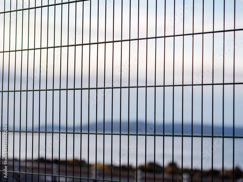 Obraz na płótnie Metal wired fence in focus, ocean rock shore and mountains in the background. Blue color and dark tones give sad imprisonment mood. Huge open nature space symbolize freedom, fence shows limitations. w salonie