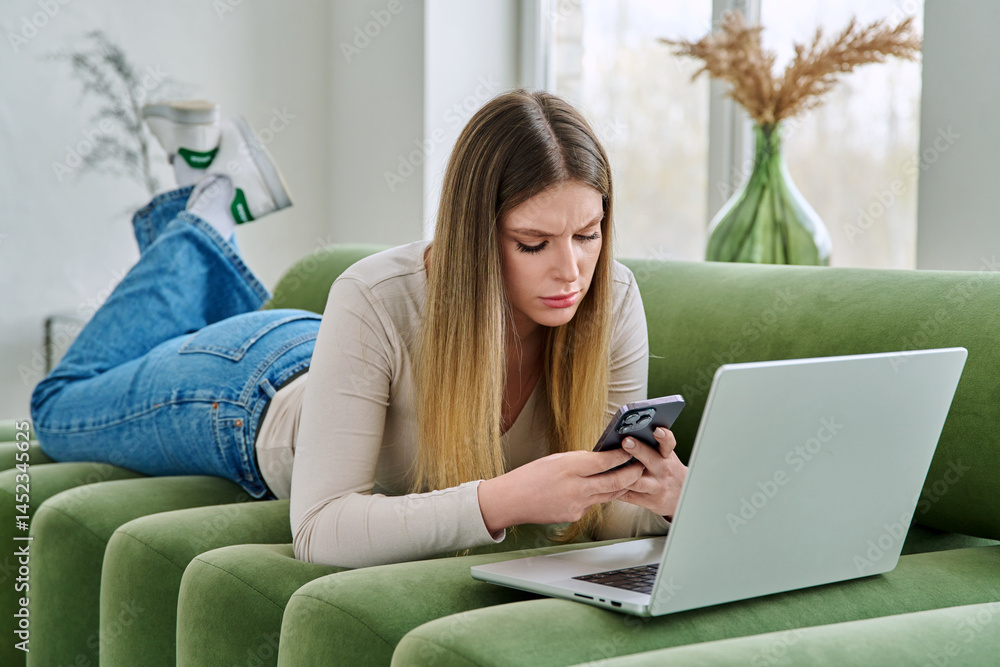 Young serious woman lying on couch with laptop, using smartphone
