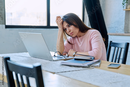 Middle aged sad stressed woman sitting at home at table with computer