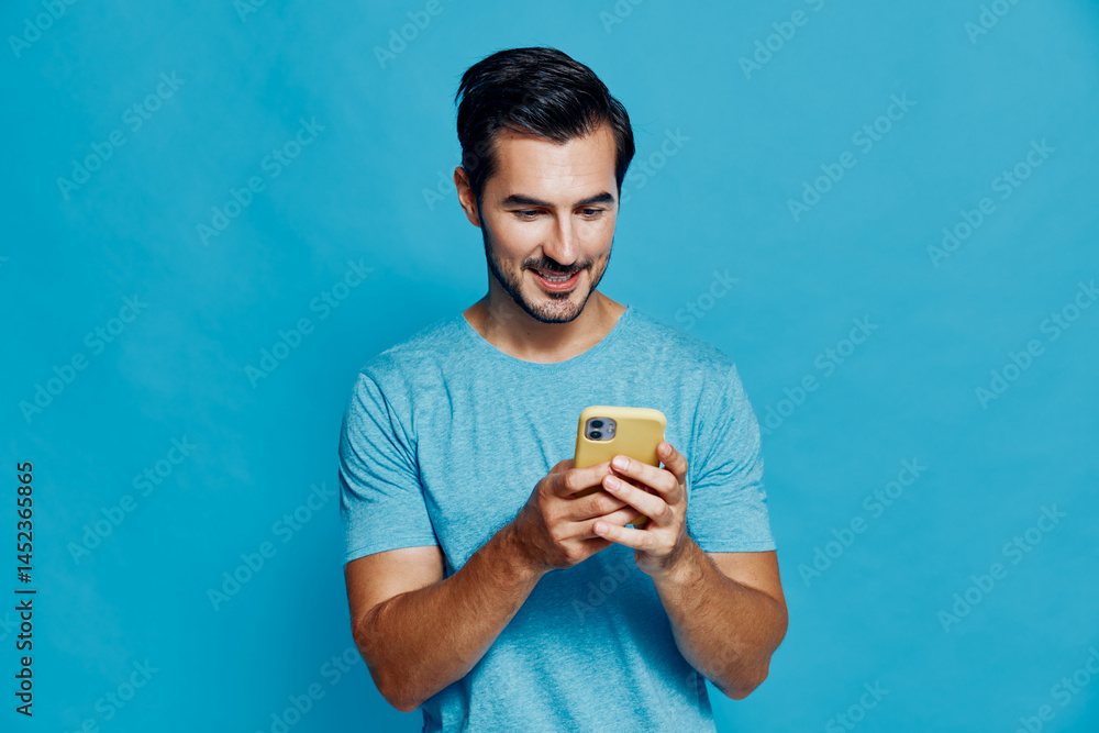 Smiling man wearing a light blue t-shirt, looking at his mobile phone with a cheerful expression against a solid blue background in a studio portrait.