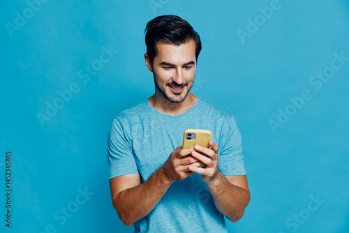 Obraz na płótnie Smiling man wearing a light blue t-shirt, looking at his mobile phone with a cheerful expression against a solid blue background in a studio portrait. w salonie