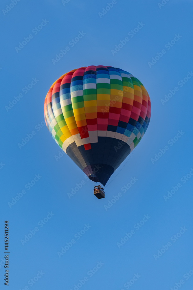 Fototapeta premium Colorful hot air balloon flying in a clear blue sky above red-tiled rooftops of residential buildings during golden hour