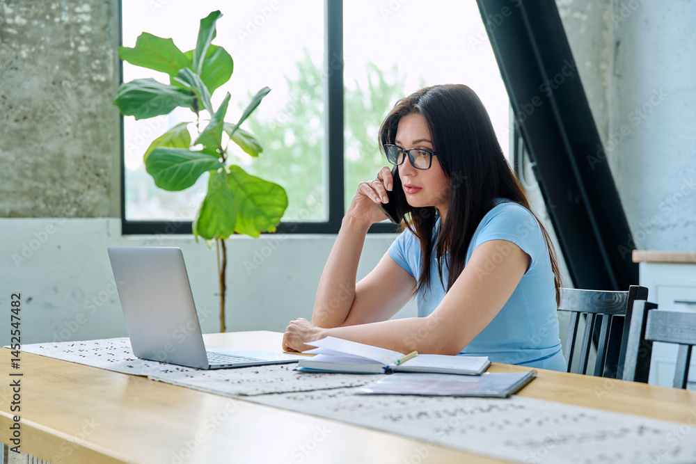 Young woman working with laptop, talking on smartphone sitting at table at home