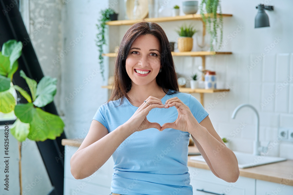 Portrait young smiling woman making heart gesture with her hands at home