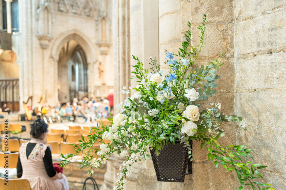 Shallow focus of an artificial flower arrangement seen on a pillar of a famous English cathedral. People can be sitting on chairs within the cathedral listing to a live orchestrator practice.