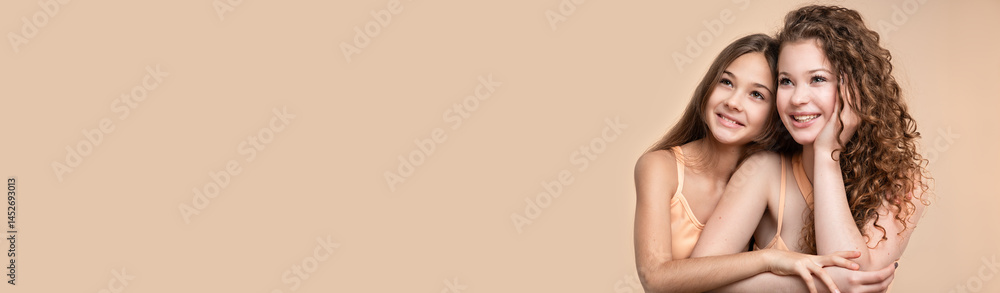 Banner with studio portrait of two beautiful smiling sisters sitting together against beige background
