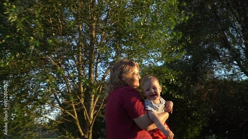 A young mother carries her one-year-old smiling son in front of her in a park at sunset in summer. Mother and son pastime