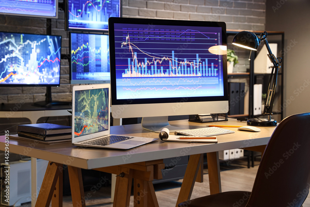 Computer and laptop with stock data on trader's desk in dark office Computer and laptop with stock data on trader's desk in dark office