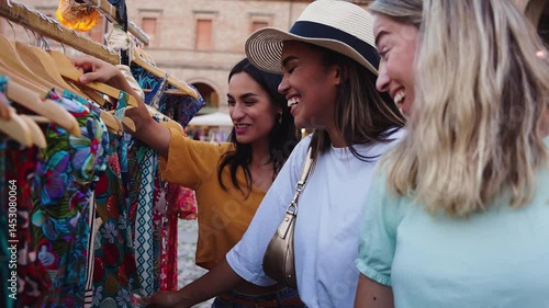Three young women are shopping for clothes at an outdoor market in piazza santo stefano in bologna, italy, enjoying their vacation and browsing through colorful garments