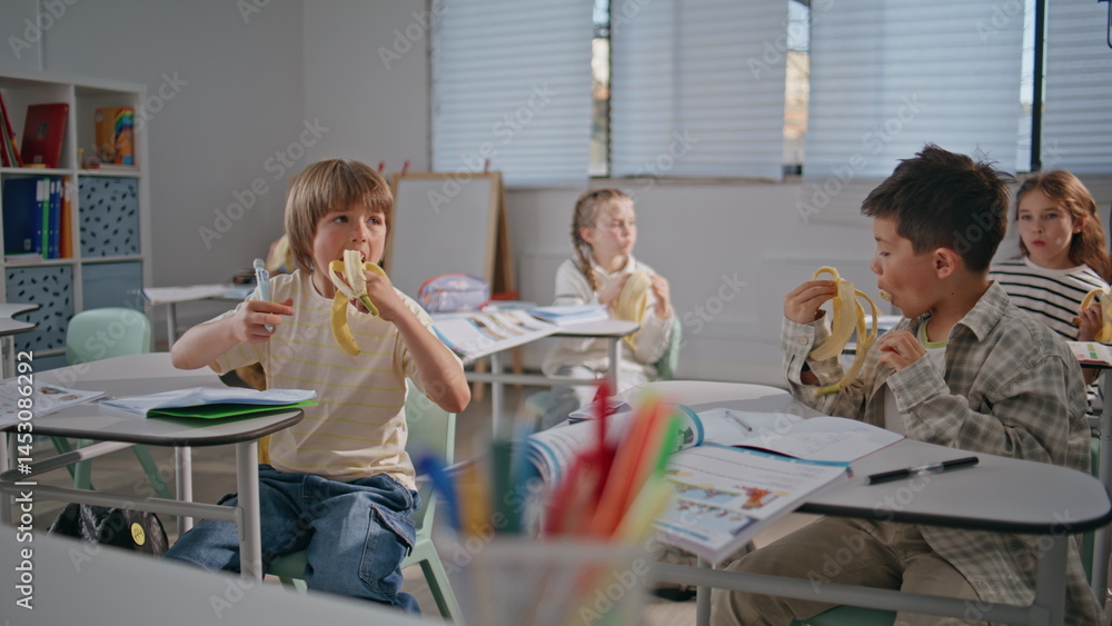 Two boys eating bananas at school desks. Hungry classmates enjoying lunch break