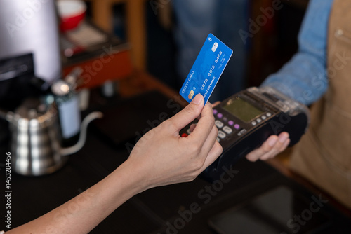 Close-up of customer hand holding blue credit card making electronic payment using point of sale terminal held by barista in small coffee shop representing modern contactless transaction service