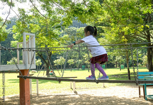 An Asian young girl wearing a white shirt, red plaid skirt, and purple knee-high socks carefully balances on a rope bridge at an outdoor playground in a lush green park